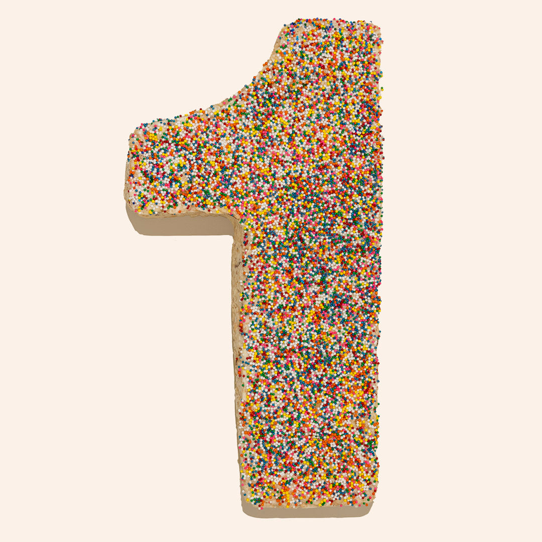 Food photography of a cake in the shape of the number one centered on a plain background featuring a dense layer of vibrant rainbow nonpareil sprinkles, thick textured cream frosting along the edges, and a soft cast shadow