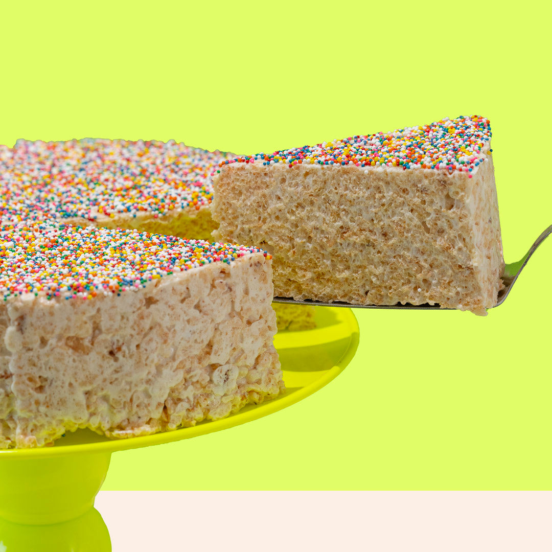 round rice cereal treat cake topped with rainbow sprinkles, with a slice being pulled out of the cake
