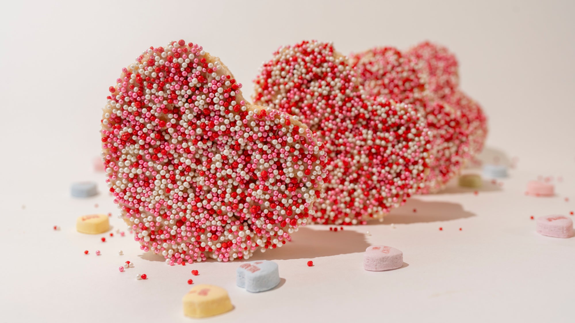 heart cut out rice cereal treat bars, coated in pink and red and white sprinkles, with conversasion hearts scattered in the foreground