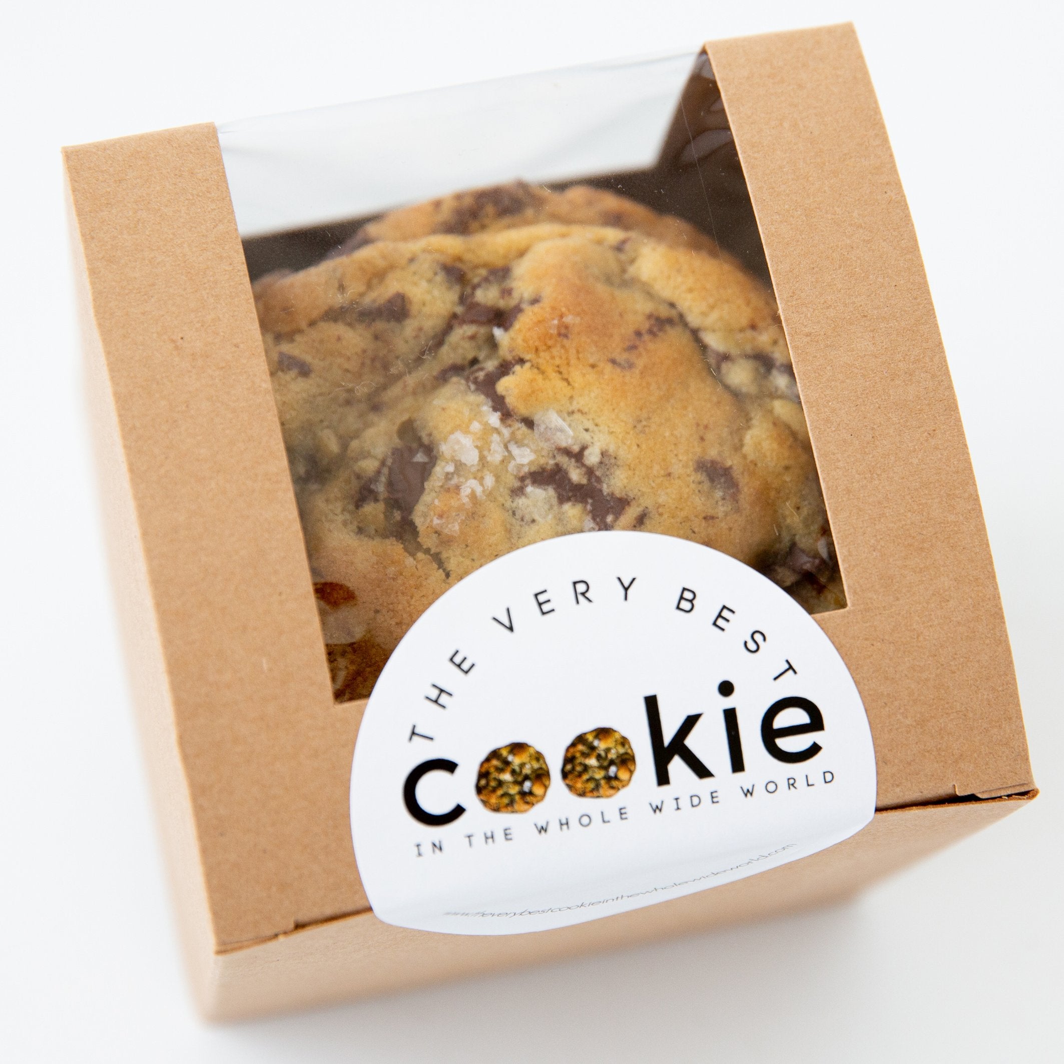 A freshly baked, gourmet chocolate chip cookie sprinkled with sea salt sits in a kraft window box, ready to be enjoyed.