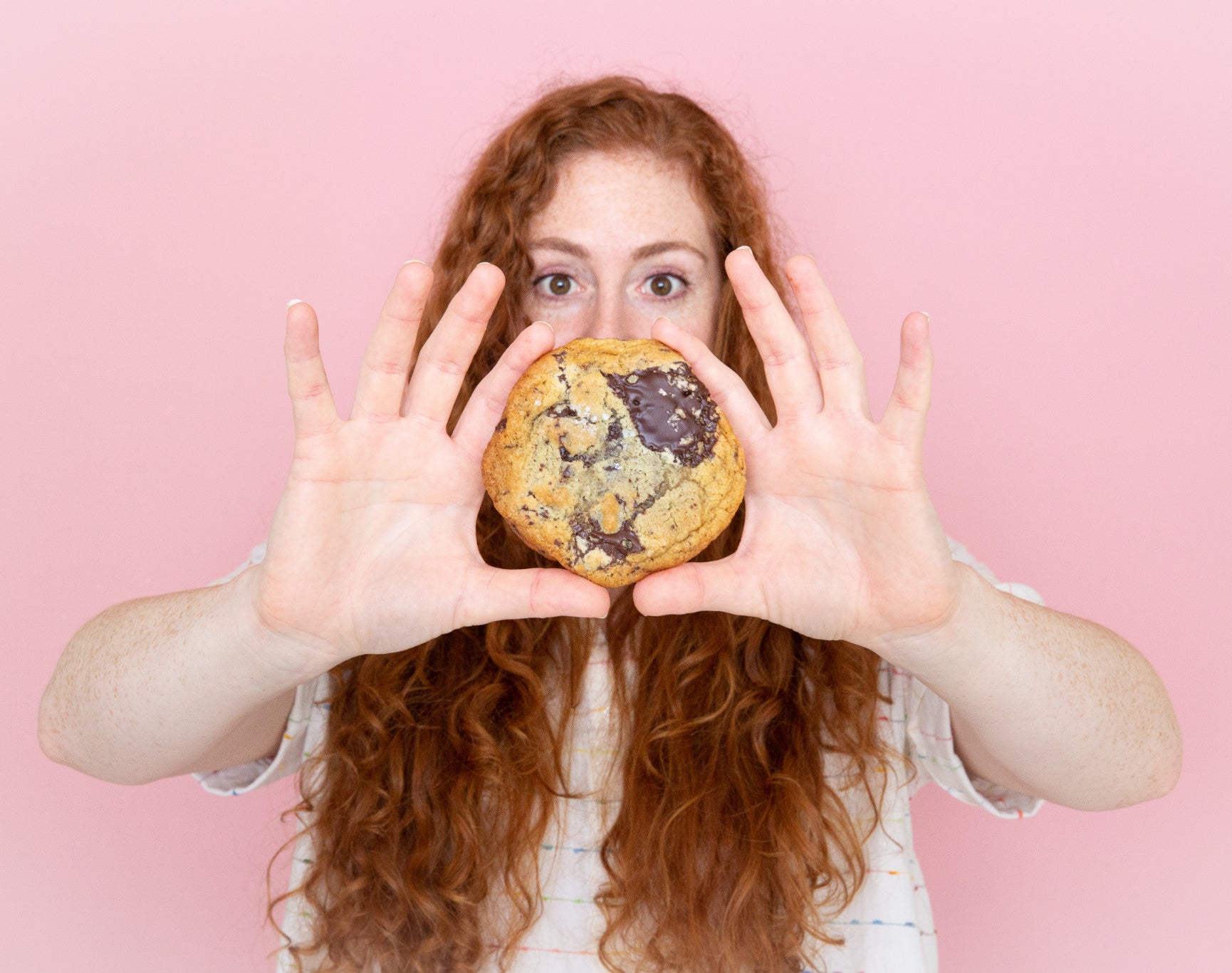Woman holds a large, freshly baked chocolate chip cookie in front of her face against a pink background. 
