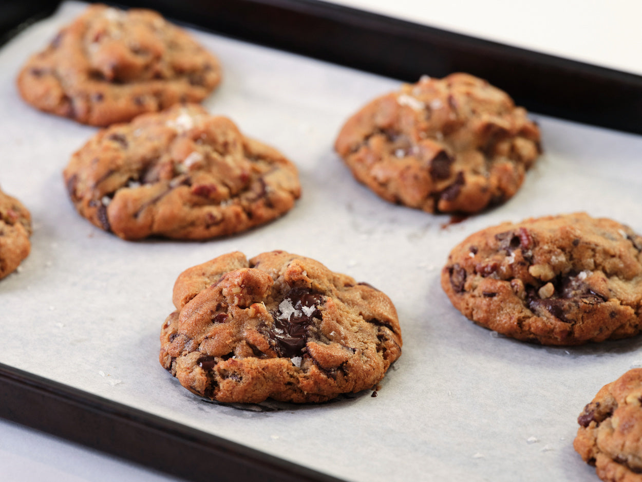 Six freshly baked chocolate chunk pecan cookies, topped with flaky sea salt, cool on a baking sheet lined with parchment paper. 
