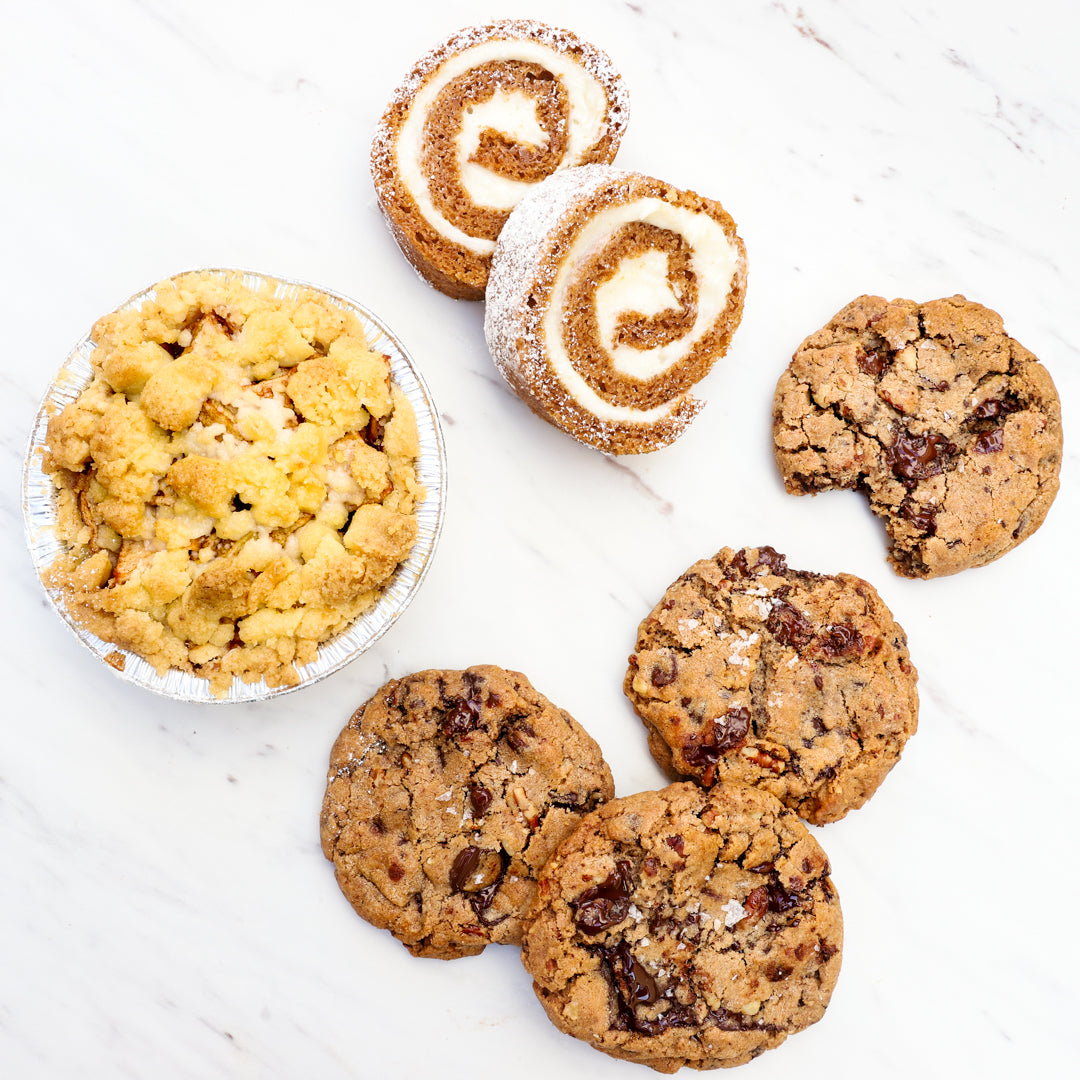 A flat lay of assorted baked goods on a white marble surface featuring a mini apple crumble pie, two cinnamon-swirl pumpkin roll slices dusted with powdered sugar, and four chocolate chip pecan cookies with sea salt flakes, one partially eaten