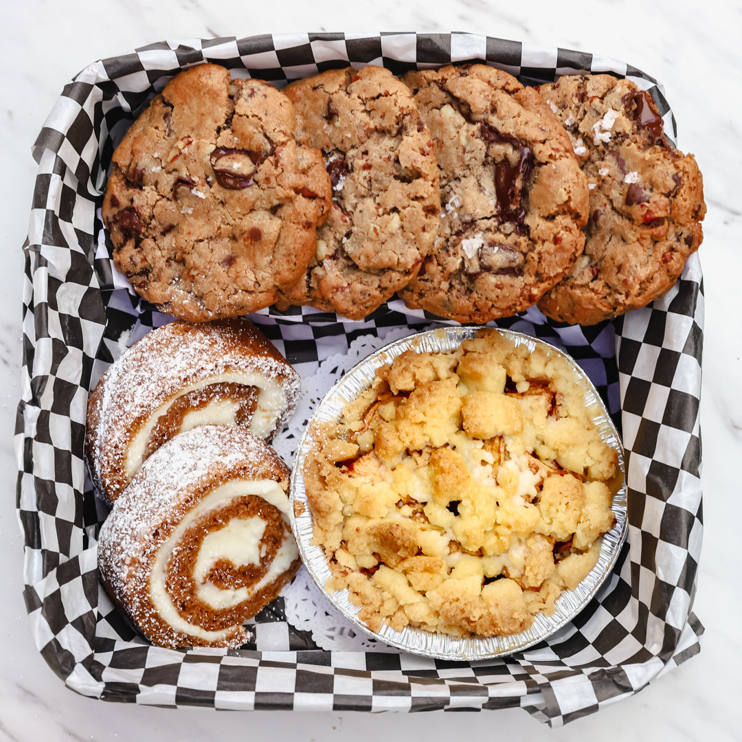 An assortment of baked goods featuring four sea salt chocolate chip cookies, two powdered sugar pumpkin rolls with cream cheese filling, and a mini apple crumble pie, arranged in a black and white checkered paper-lined basket on a white marble background